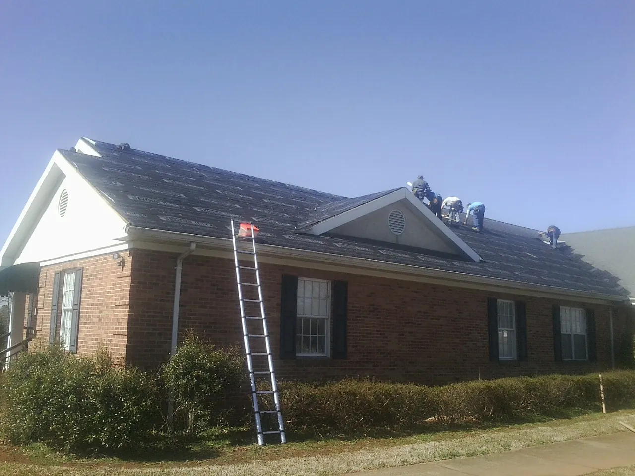 Crew tearing shingles off a brick ranch roof with a ladder set — Emergency Roof Repair work by our crew in Tigerton