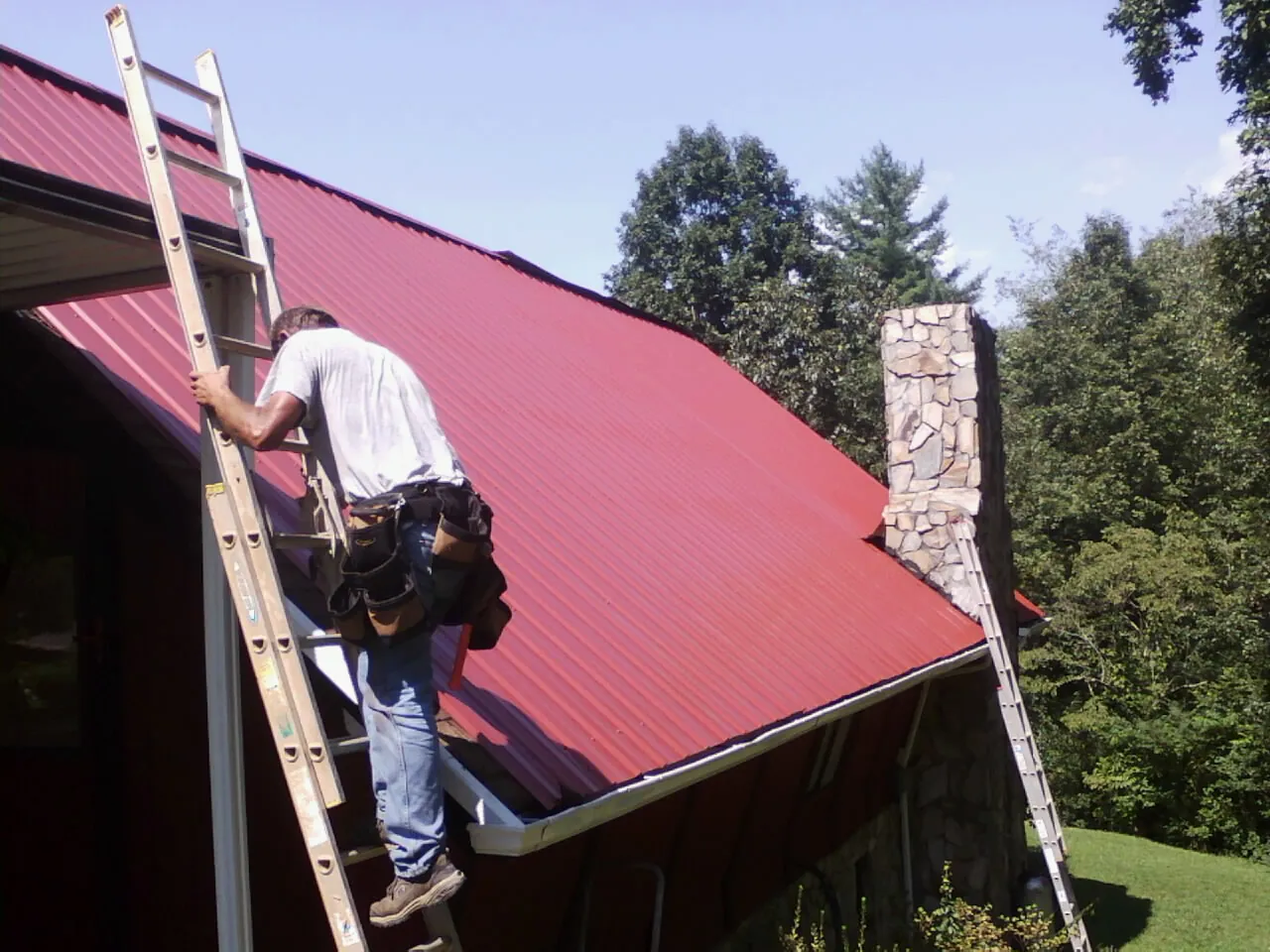 Roofer on a ladder beside a red metal roof and stone chimney — Soffit & Fascia Repair by Roofing Brothers in Tigerton